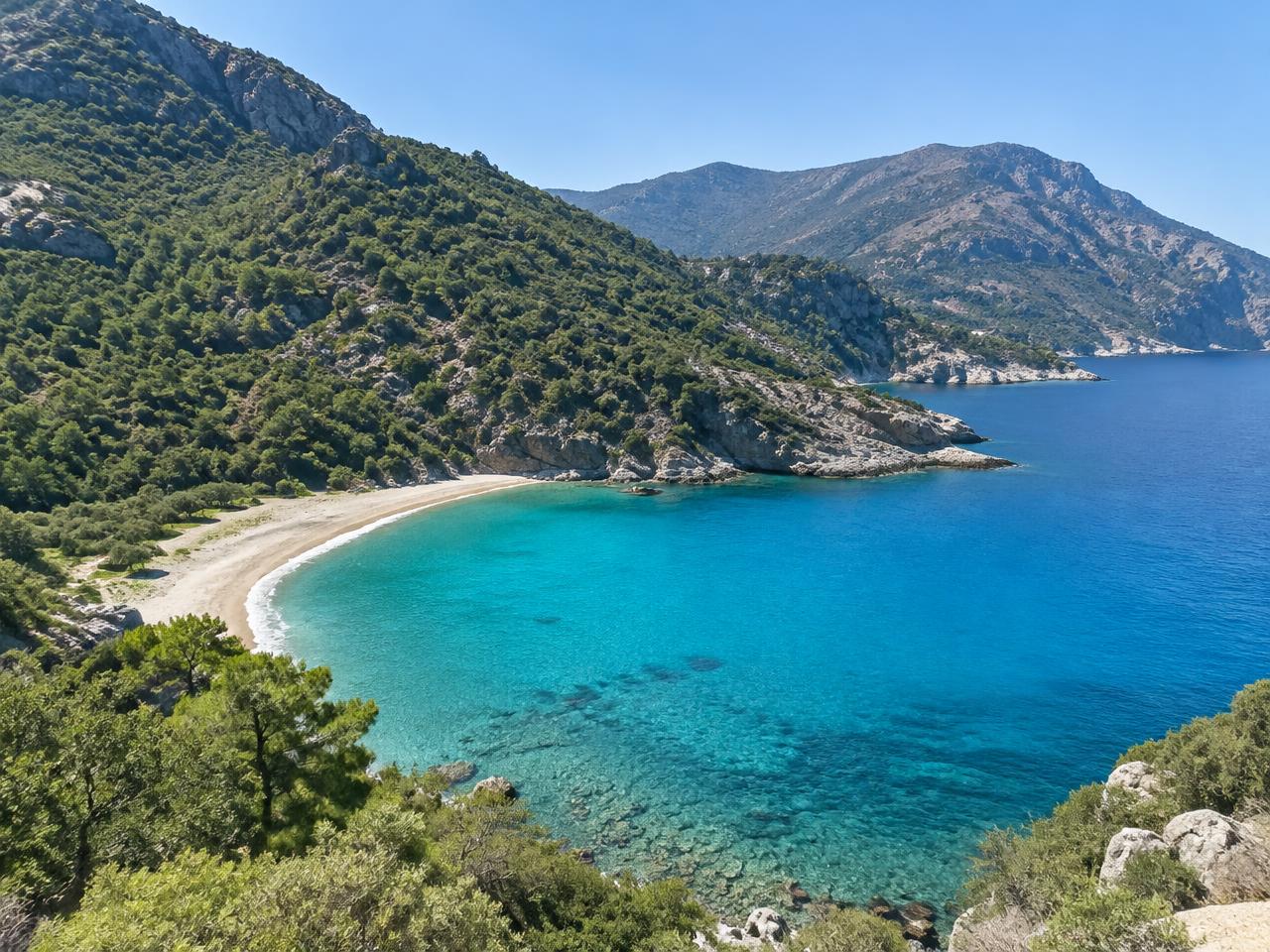 Bewaldete Berghänge über einer Sandbucht mit klarem Wasser auf Ikaria