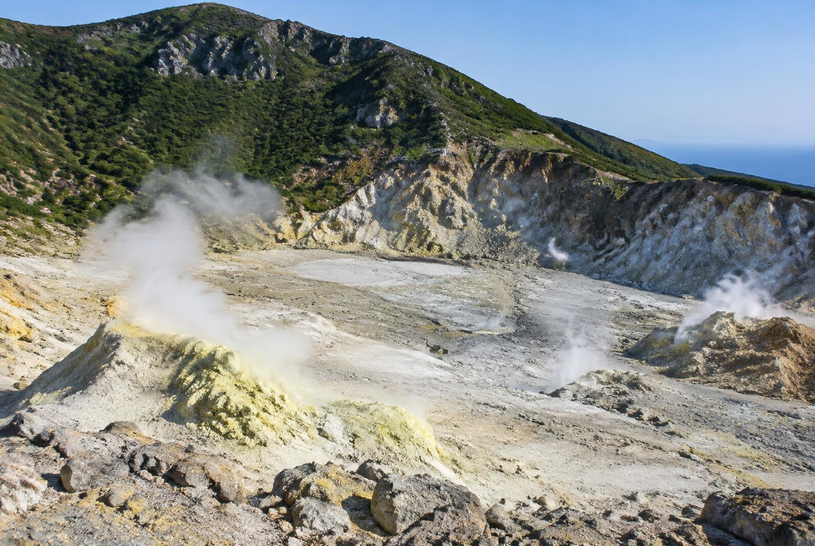 Vulkankrater mit hellem, dampfendem Boden, dahinter grüne Kraterhänge